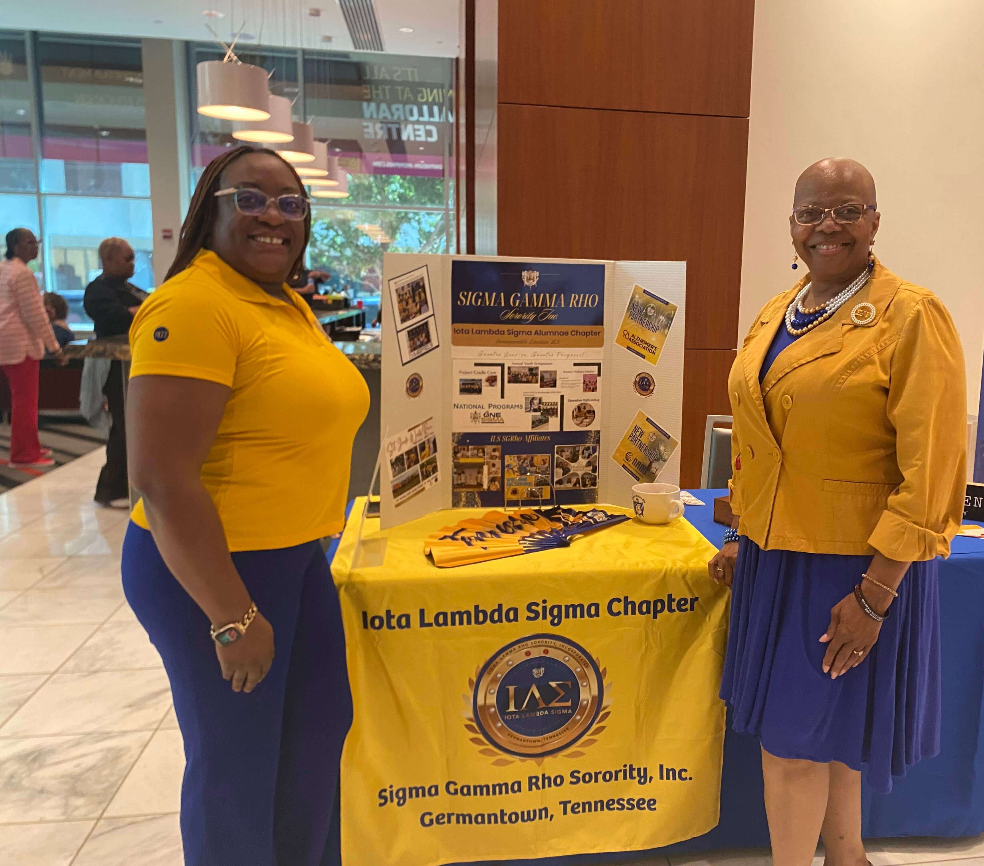 Members of Sigma Gamma Rho Sorority stand beside an informational display and table promoting community programs and partnerships at an event