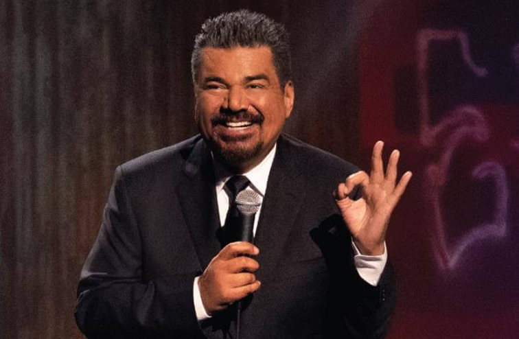 George Lopez on stage holding a microphone and wearing a suit for the filming of his Netflix special, We'll do it for half.