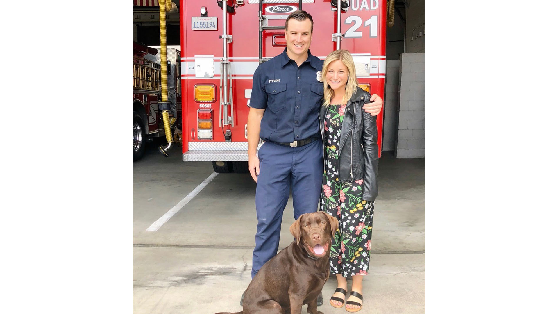 Firefighter Eric Stevens and his wife pose with their dog in front of a fire engine at a fire station.
