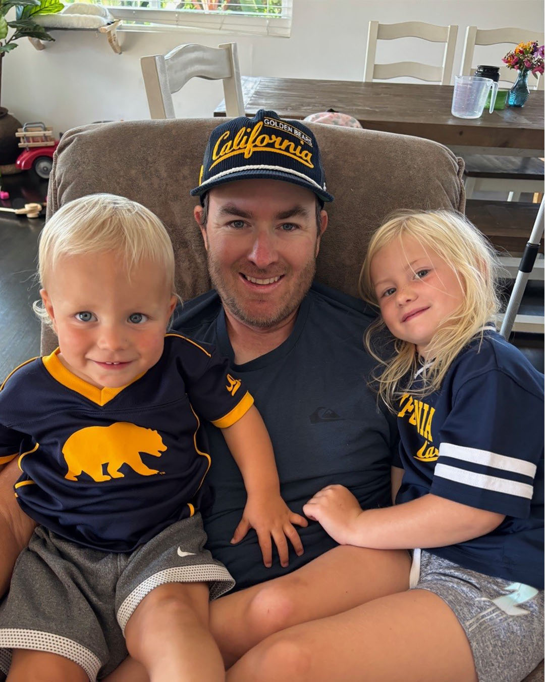 Eric Stevens sits with his two young children at home, all wearing University of California Golden Bears apparel and smiling.