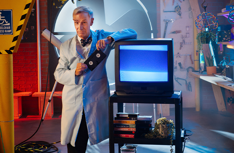 Bill Nye holding up a VHS tape and leaning against a cart holding a TV