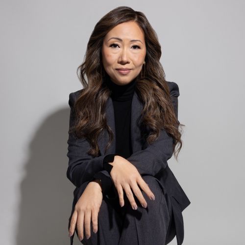 Dr. Fong-Isariyawongse sitting down with her arms crossed over each other, wearing a dark gray suit and black top with curled hair and closed mouth smiling