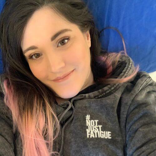 Elizabeth Ansell, founder of #NotJustFatigue, smiling while lying down and wearing a dark hoodie with the hashtag “#NotJustFatigue” printed on it.