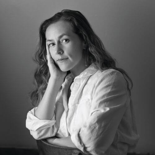Portrait of Greta Morgan, seated and looking thoughtfully at the camera, wearing a light-colored shirt with long, wavy hair.