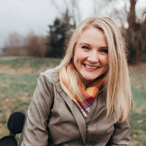 Smiling woman with long blonde hair wearing a colorful scarf and olive green jacket, sitting outdoors on a cloudy day
