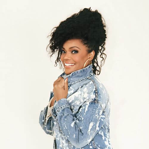 Yvette Nicole Brown smiling and looking over her shoulder while wearing a splatter-painted denim jacket, against a white background.