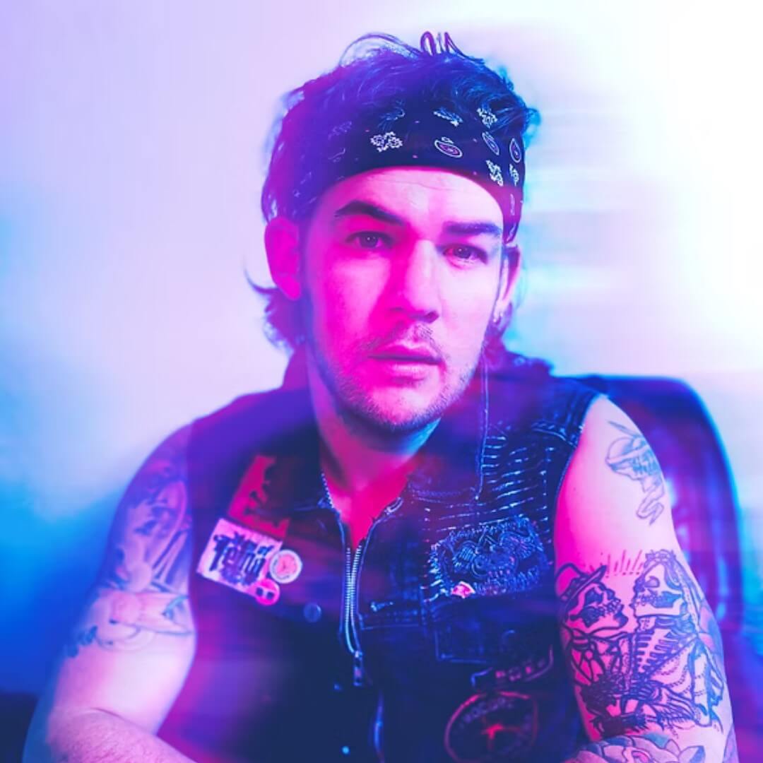 James Durbin wearing a sleeveless denim vest and bandana, with tattooed arms, photographed under colorful stage lighting.